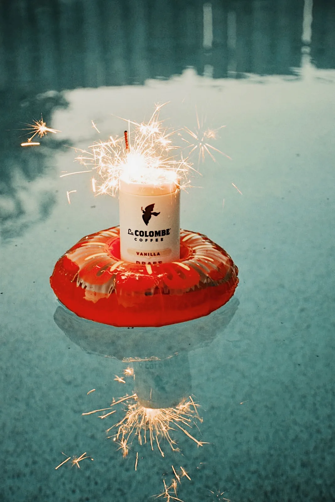 Vanilla Draft Latte can floating on a small red pool ring with a lit sparkler, reflected in the water