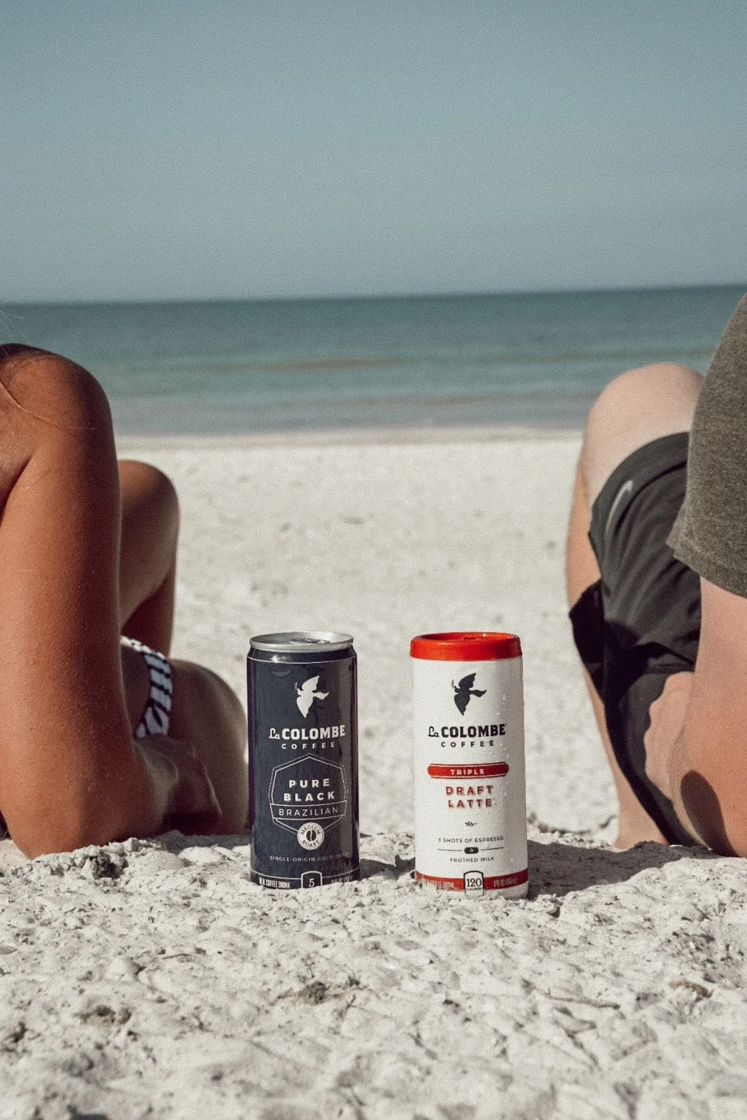 Pure Black Brazilian cold brew and Triple Draft Latte cans standing in white sand with the Gulf behind them