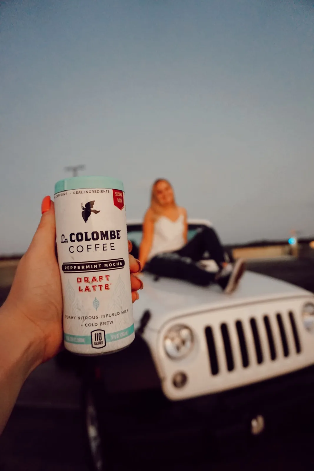Peppermint Mocha Draft Latte held in front of a white Jeep with a model seated on the hood at sunset