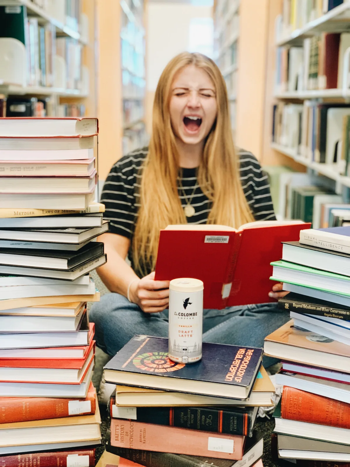 Student screaming behind towering stacks of library books with a Vanilla Draft Latte can in the foreground