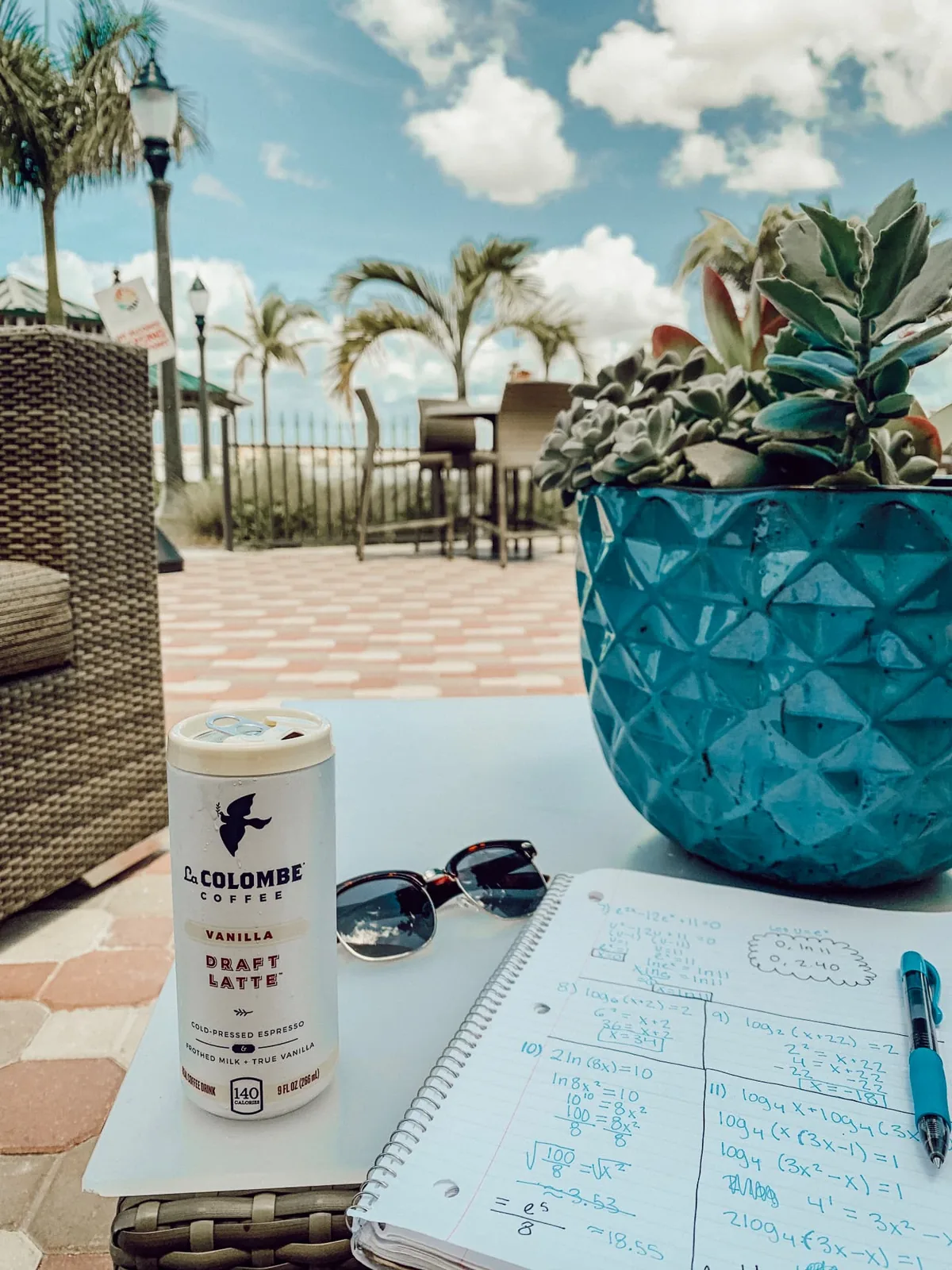 Vanilla Draft Latte on a poolside table beside sunglasses and an open notebook of calculus work, palm trees behind