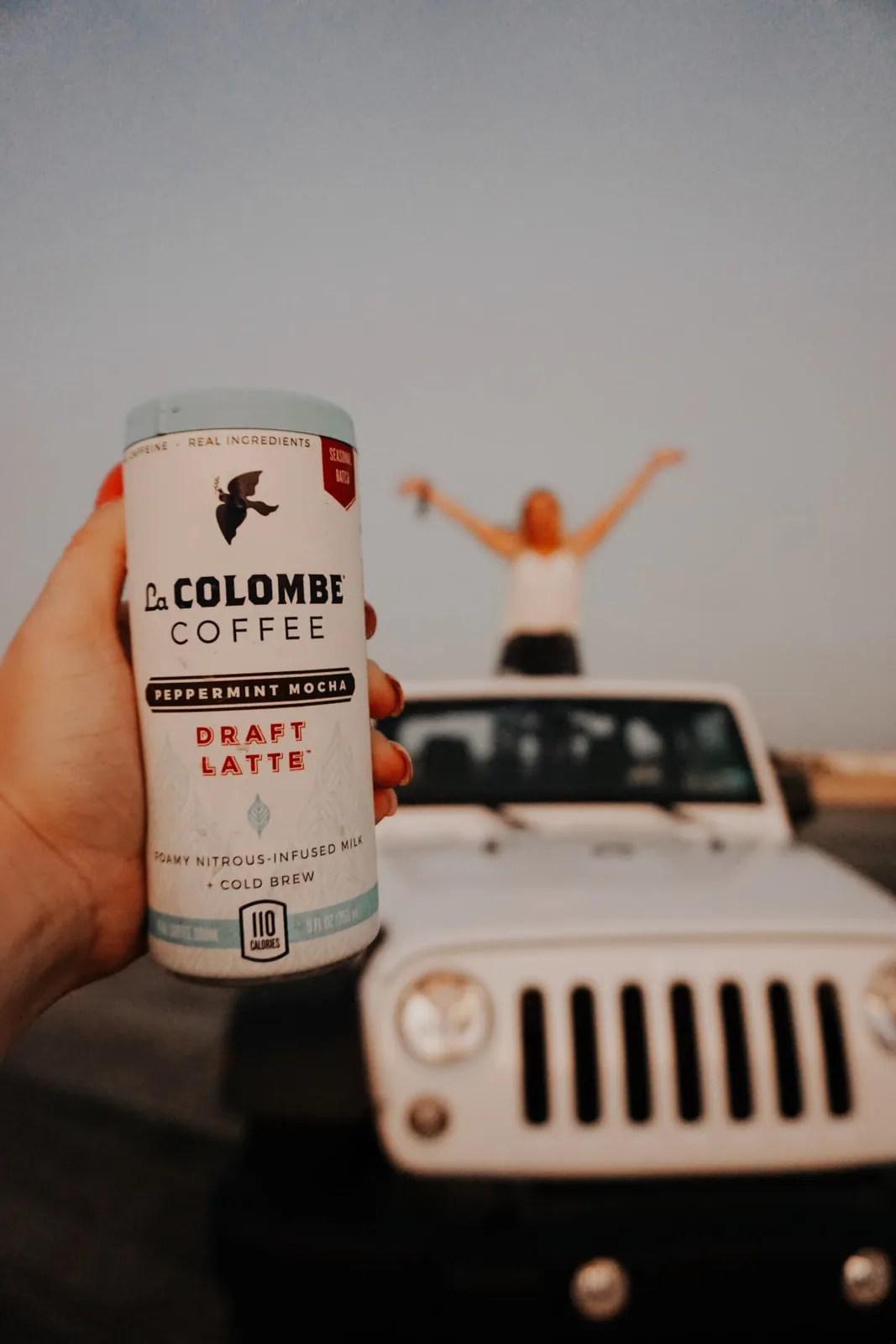 Hand holding a Peppermint Mocha Draft Latte with a person cheering on a white Jeep at dusk behind it
